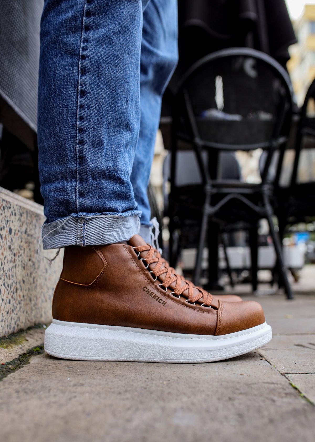 A person wearing brown high-top sneakers with white soles and laced up, standing on a stone surface with black chairs and a table in the background.