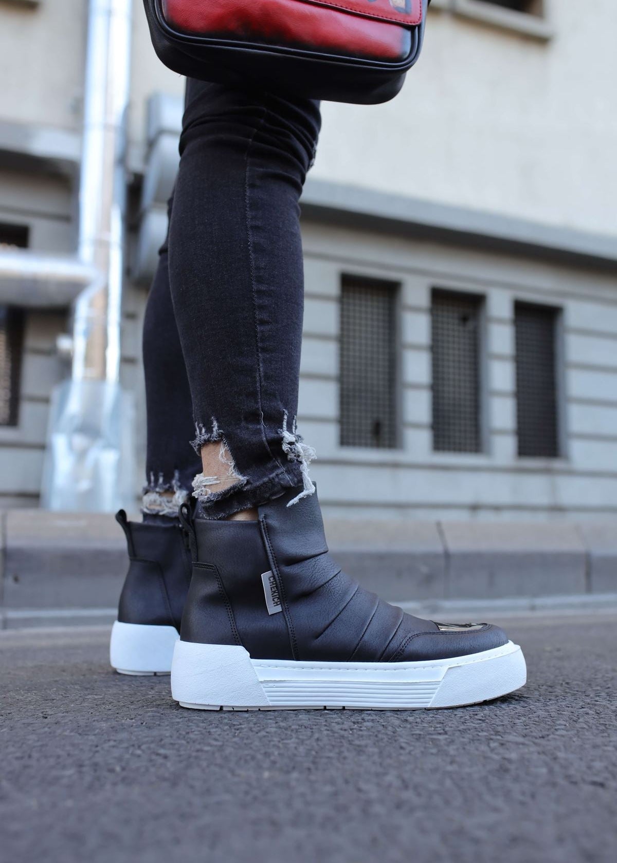 A pair of men's boots with black vegan leather upper, white sole, and a mirror toe cap. The boots are displayed with a person's jeans and a casual top, showcasing the sneaker-style zipper and the brand logo on the side.