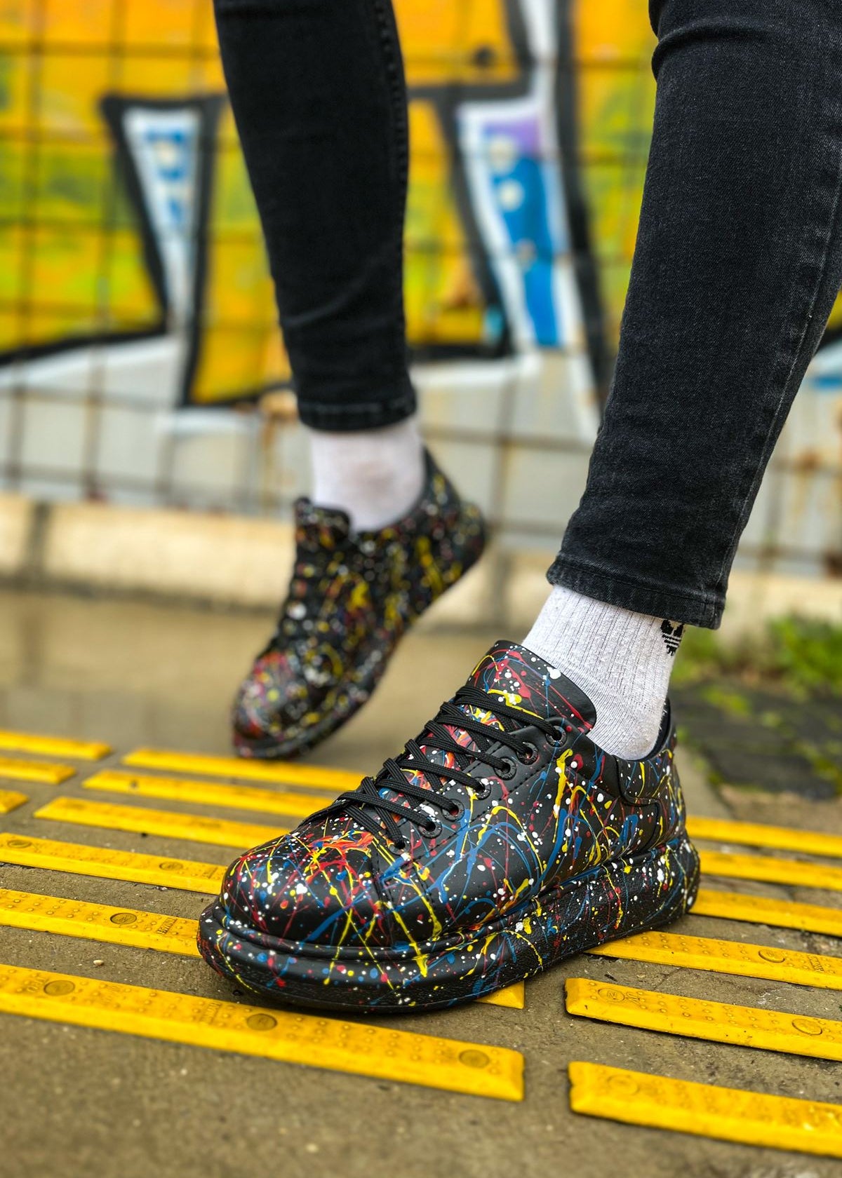Colorful sneakers with black soles worn on a yellow tactile paving surface.