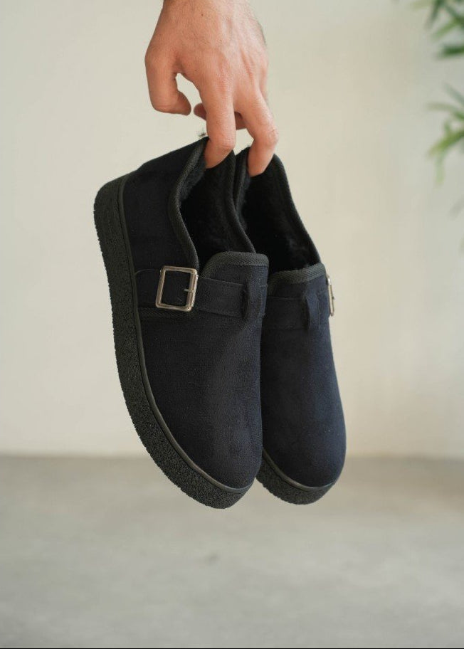 Black slippers with a buckle held by a hand against a neutral background