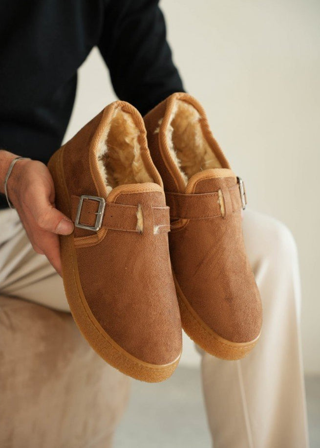 Brown suede slippers with fur lining held by a person against a neutral background